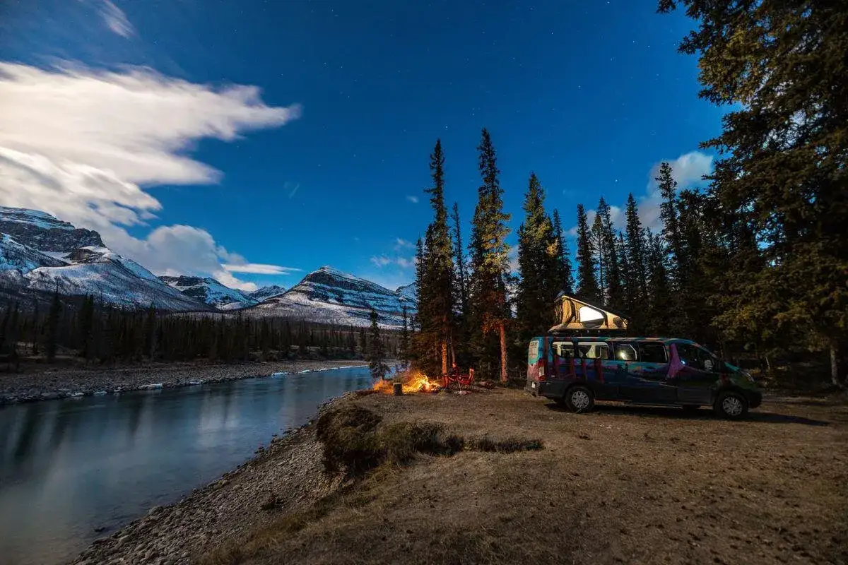 Un camping-car coloré pour un road trip au Canada, campant près d'une rivière sous un ciel étoilé, avec des montagnes enneigées en arrière-plan.
