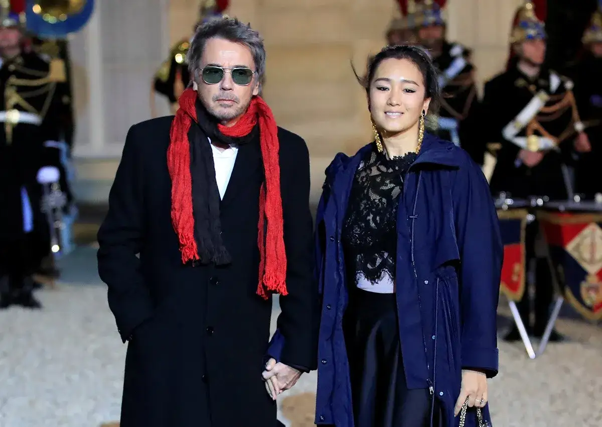 Jean-Michel Jarre z żoną, w eleganckich strojach, idą trzymając się za ręce. W tle orkiestra wojskowa.