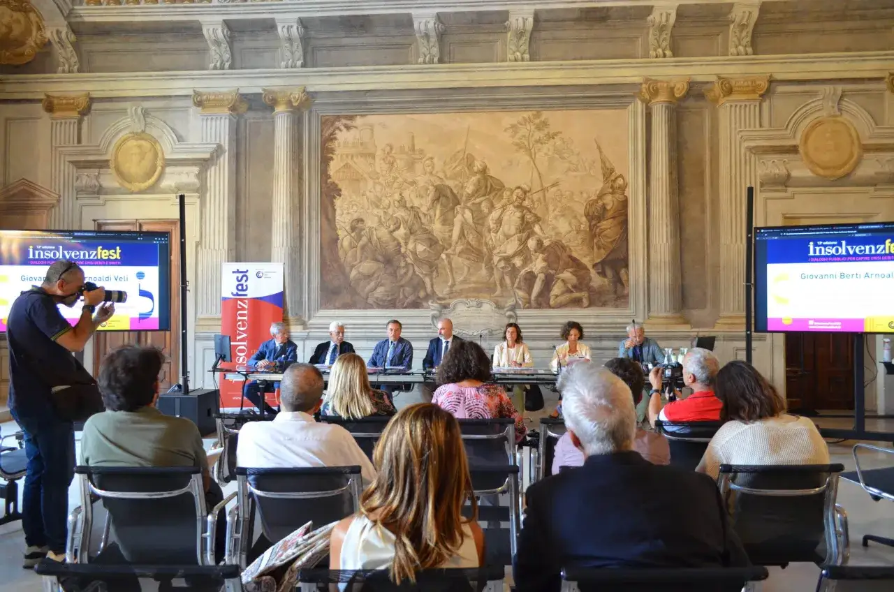 Il responsabile comunicazione documenta un convegno con relatori e pubblico in una sala affrescata.