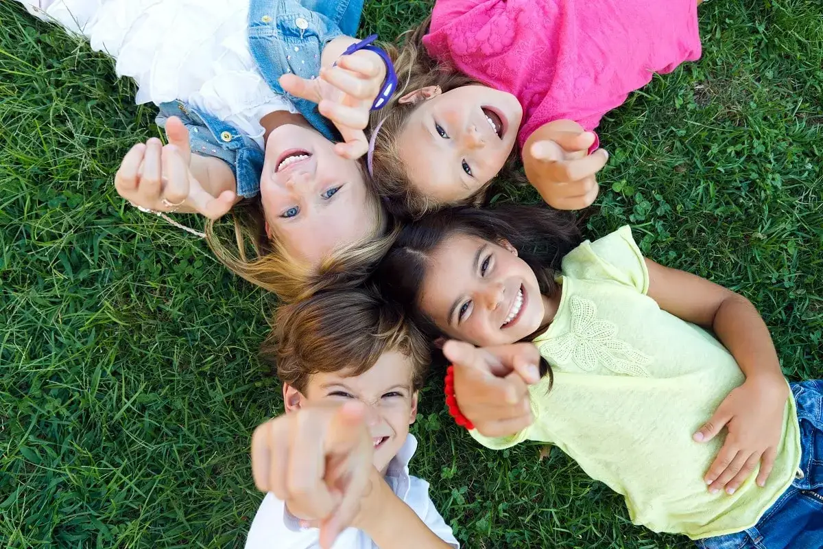 Niños sonriendo y señalando a la cámara, tumbados en el césped. ¡Perfectos para la escuela de verano de niños de 2 años!