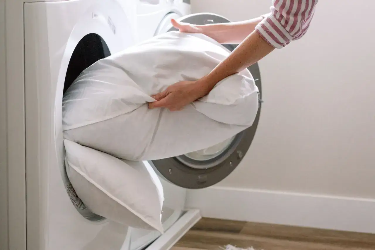 A person is placing white pillows into a washing machine, demonstrating how to wash pillows in a washing machine for a fresh, clean sleep.