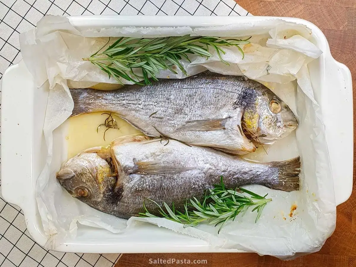 Dwie dorady w naczyniu żaroodpornym, gotowe do pieczenia, ozdobione gałązkami rozmarynu.