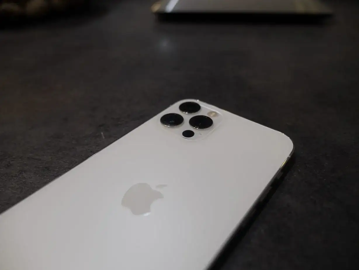 Biały iPhone 12 Pro Max z potr&oacute;jnym aparatem na ciemnym tle.