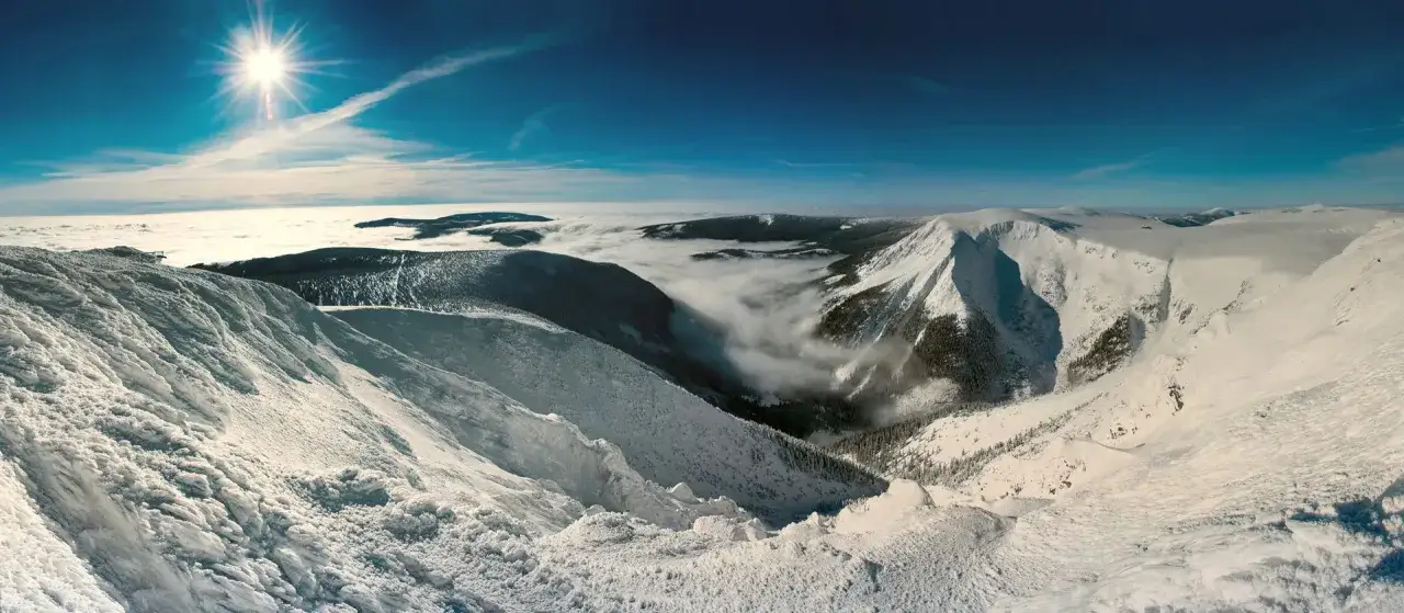 Schneebedeckte Berge in Tschechien im Winter, mit Nebel, der sich durch die Täler schlängelt und die Sonne scheint hell am blauen Himmel.