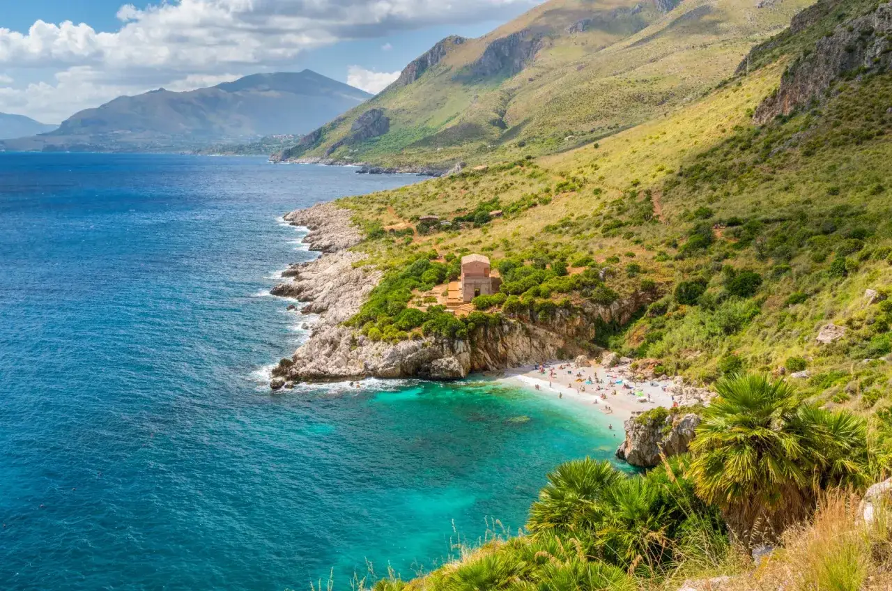 Spiagge libere Sicilia: una baia incantevole con acque turchesi, gente che prende il sole e un edificio storico su una scogliera.