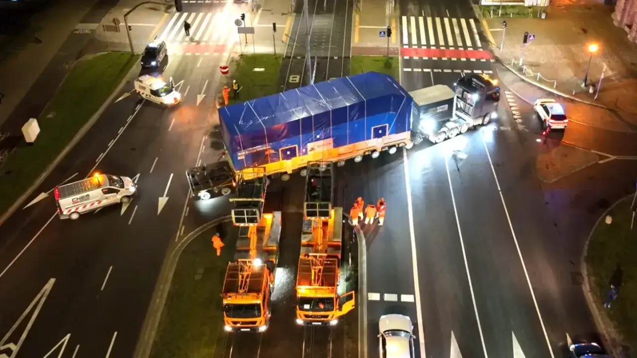Przewożąc ładunek gabarytowy, możesz poruszać się tylko z pomocą pilotów i specjalistycznych pojazdów. Ciężarówka z ogromnym ładunkiem na przyczepie, otoczona przez pojazdy pilotujące i pracowników w kamizelkach.