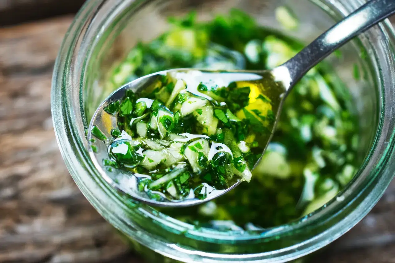 Łyżka pełna aromatycznego sosu chimichurri, z widocznymi kawałkami ziół i czosnku, zanurzona w słoiku.