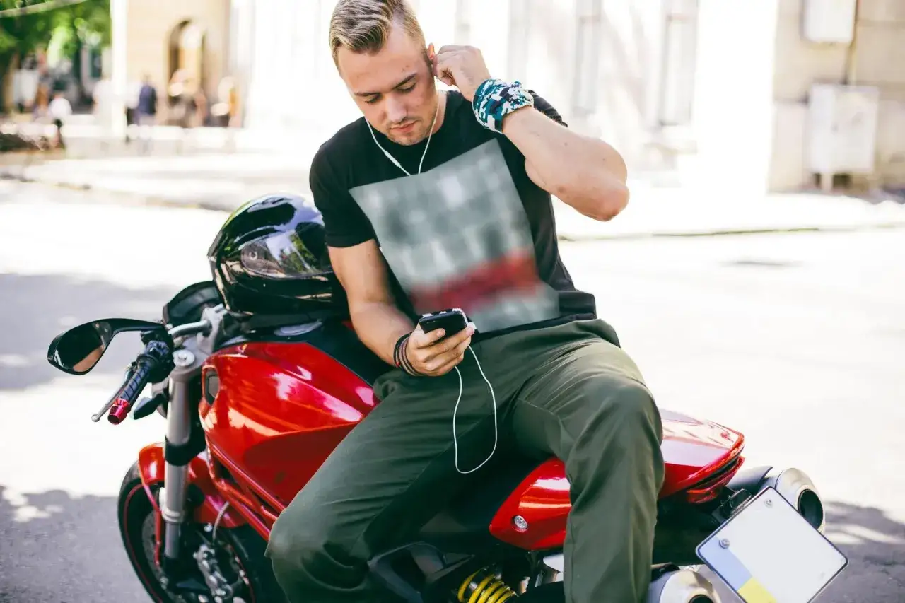 Joven en moto roja ajusta sus auriculares, disfrutando de música. ¿Es legal escuchar música en moto? ¡Él parece que sí!