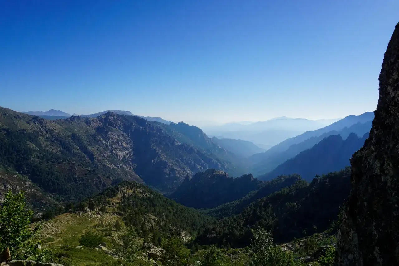 Vasta valle montuosa con cime rocciose e fitte foreste di pini. In lontananza, il cielo azzurro sfuma in una foschia leggera, creando un effetto di profondit&agrave;. Il **monte rotondo** domina il paesaggio.