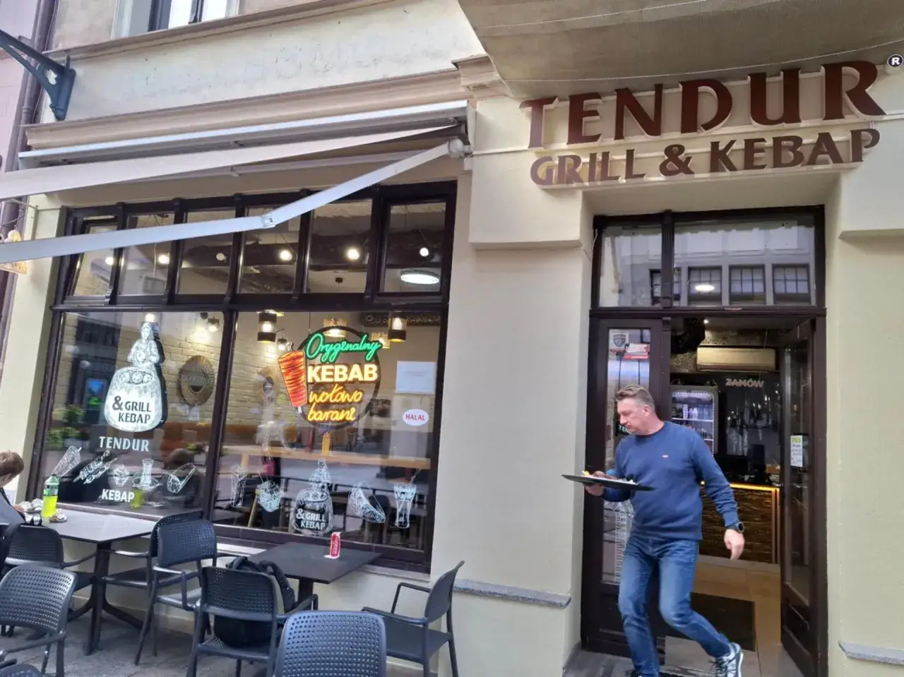 Mężczyzna wychodzi z lokalu Tendur Grill & Kebap z tacą. Na witrynie neonowy napis 