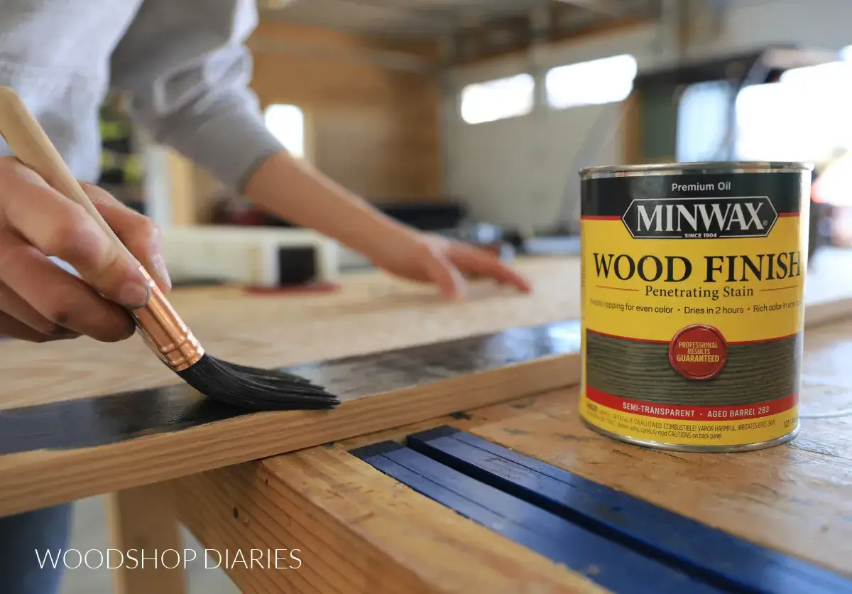 Mano che applica una macchia scura su legno con un pennello. Una lattina di Minwax Wood Finish è accanto, suggerendo che si può verniciare sopra l'impregnante.