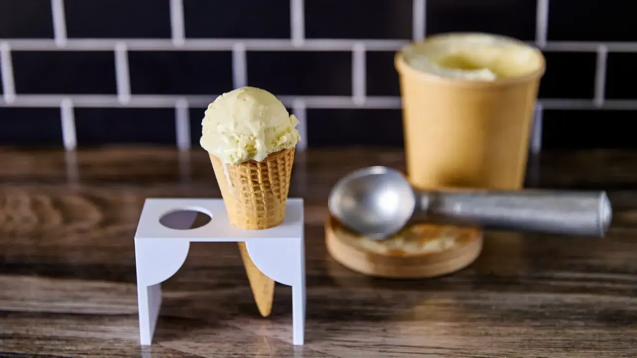 Un cono de helado de chocolate blanco cremoso, listo para disfrutar. A su lado, un cuchar&oacute;n y un recipiente con m&aacute;s helado.