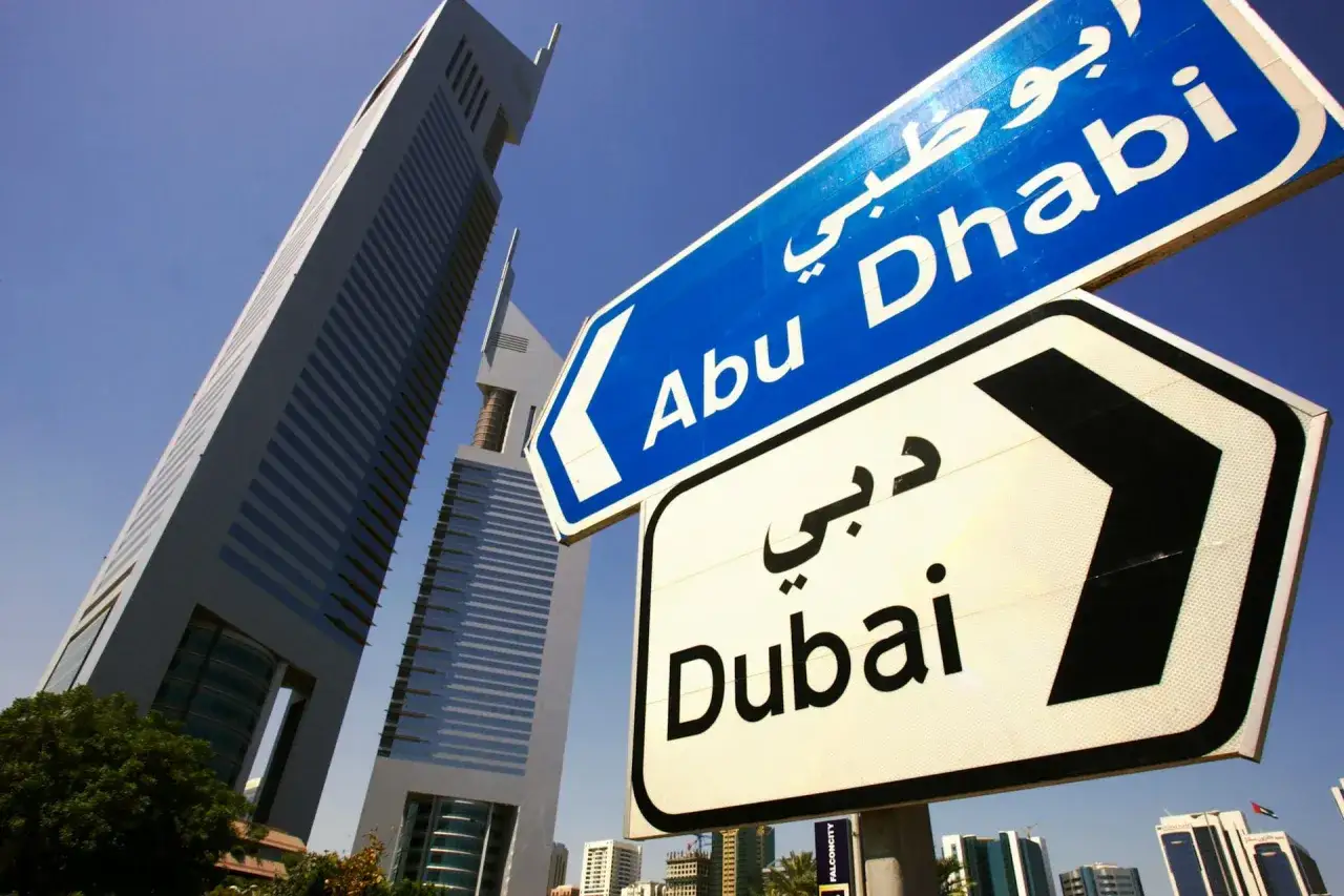 Znaki drogowe wskazują kierunek do Abu Zabi i Dubaju. W tle nowoczesne wieżowce.