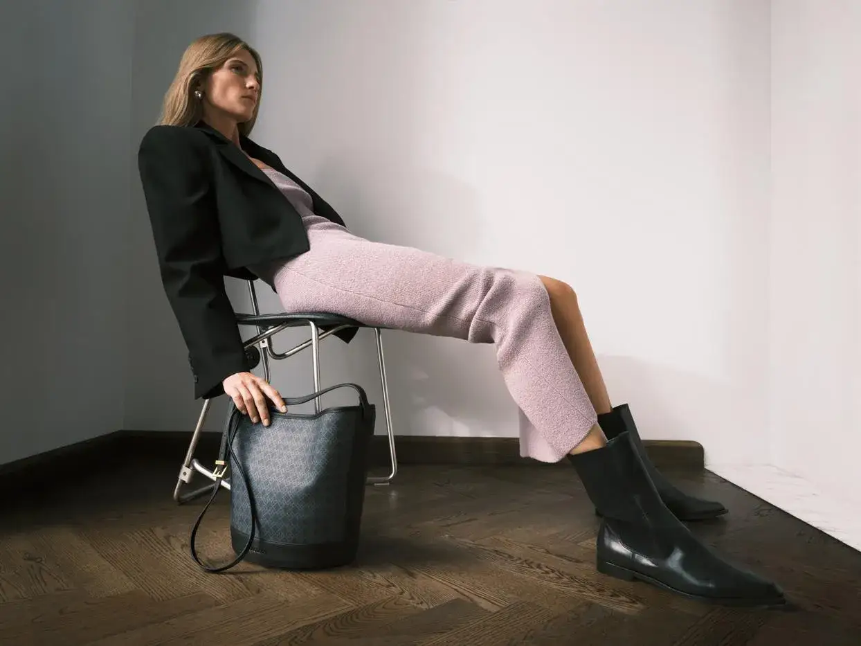 Elegancka kobieta w r&oacute;żowej sukience i czarnej marynarce, siedząca na krześle, prezentuje stylowe sztyblety.