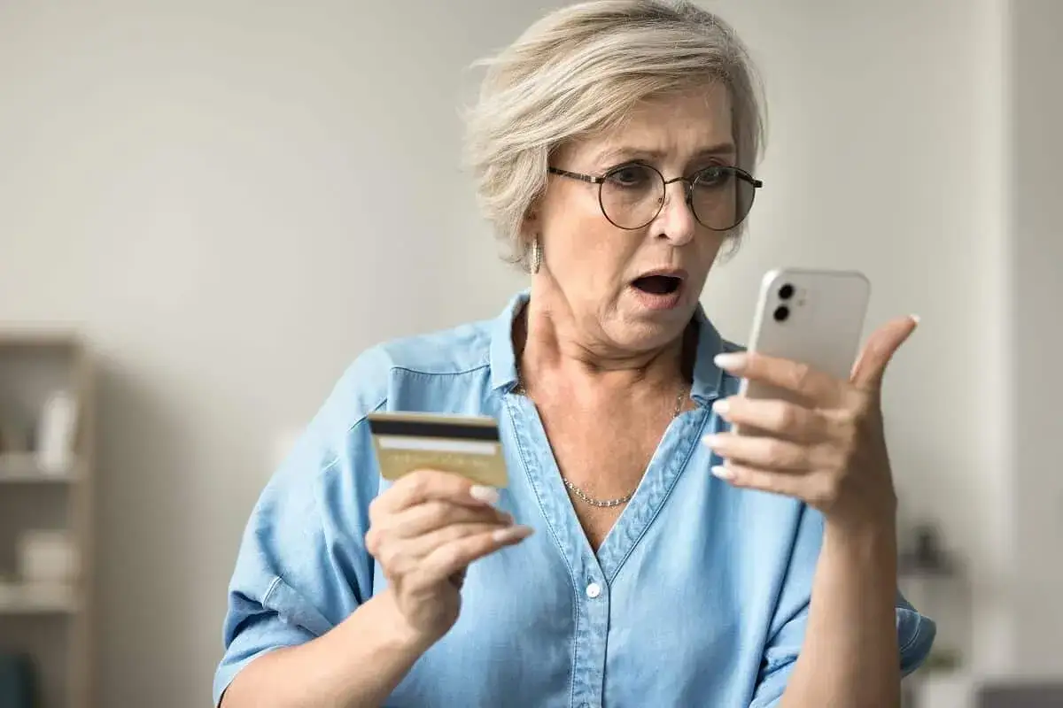 Zszokowana kobieta z kartą płatniczą w dłoni patrzy na telefon, widząc oszustwo na blik.