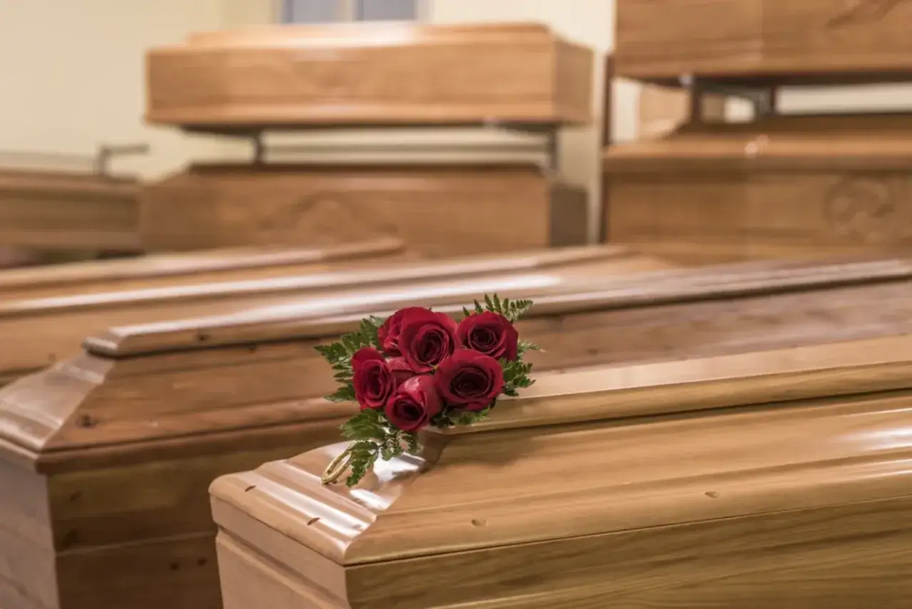 Wiele rodzajów trumien, niektóre piętrowo ułożone. Na jednej z nich bukiet czerwonych róż i złota obręcz.