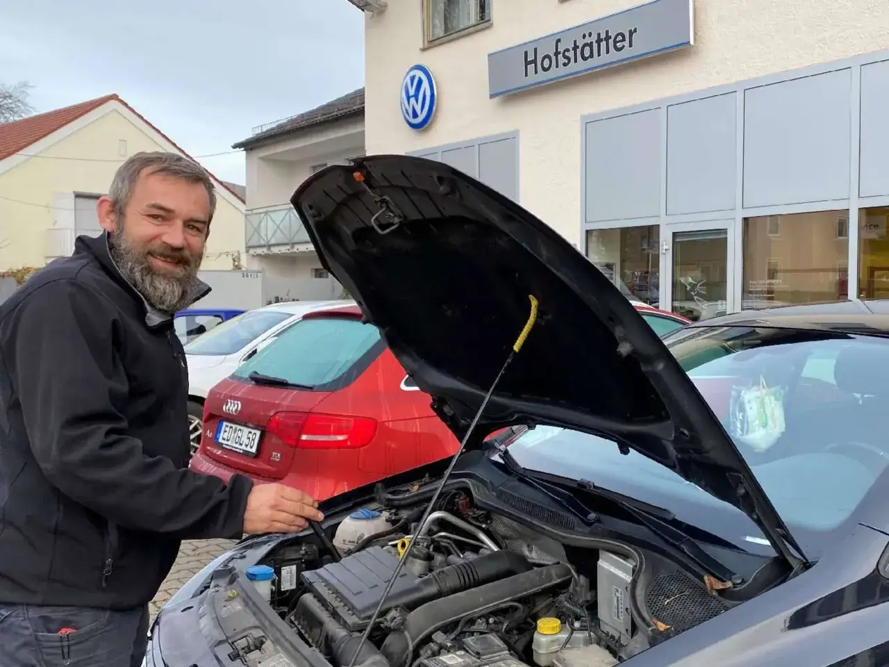 Ein Mann mit Bart steht vor einem Auto mit geöffneter Motorhaube. Im Hintergrund ist die Fassade eines Autohauses mit VW-Logo und dem Schriftzug