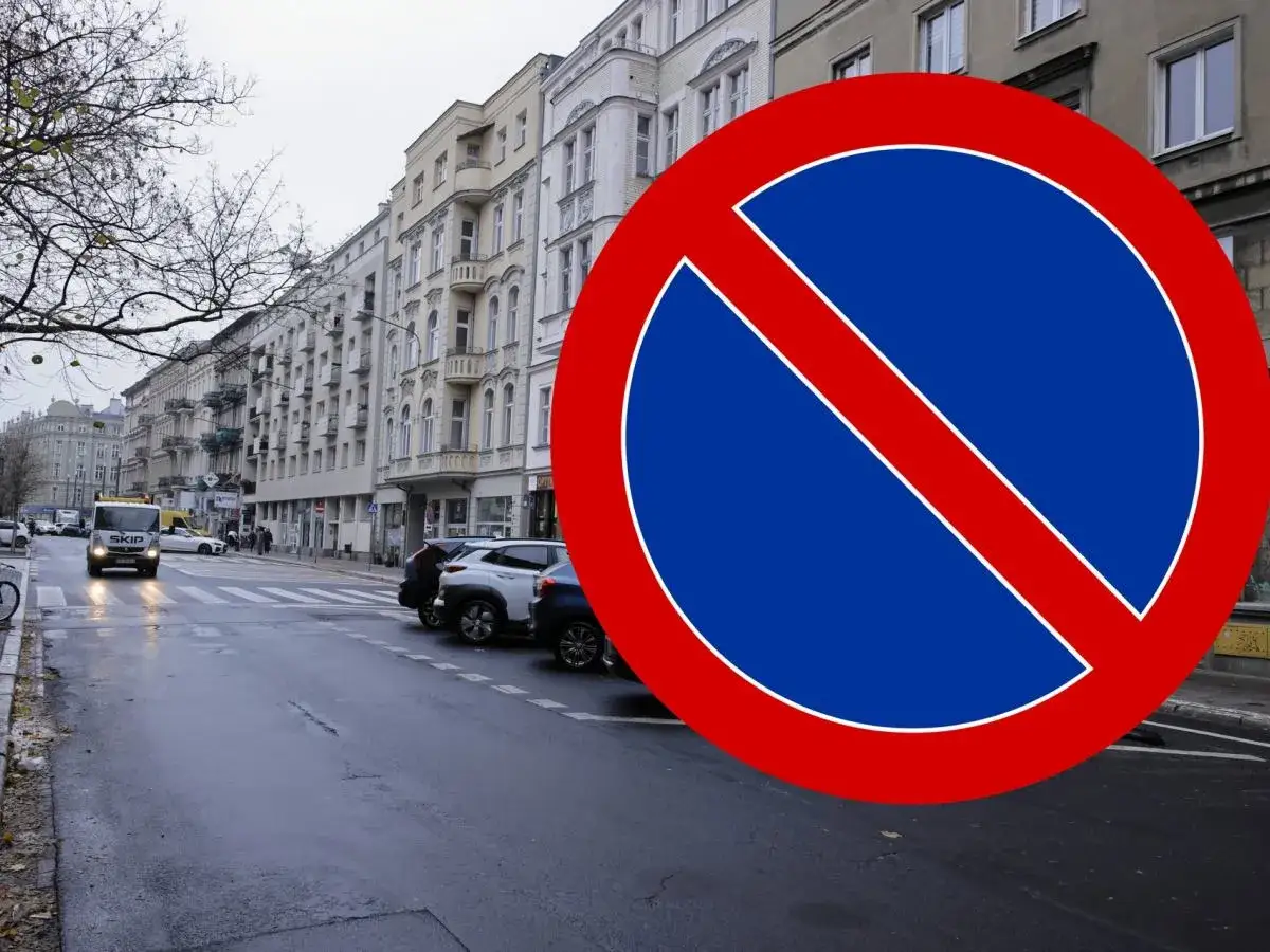 Znak B35 informuje o zakazie zatrzymywania się. Ulica z kamienicami i zaparkowanymi samochodami.