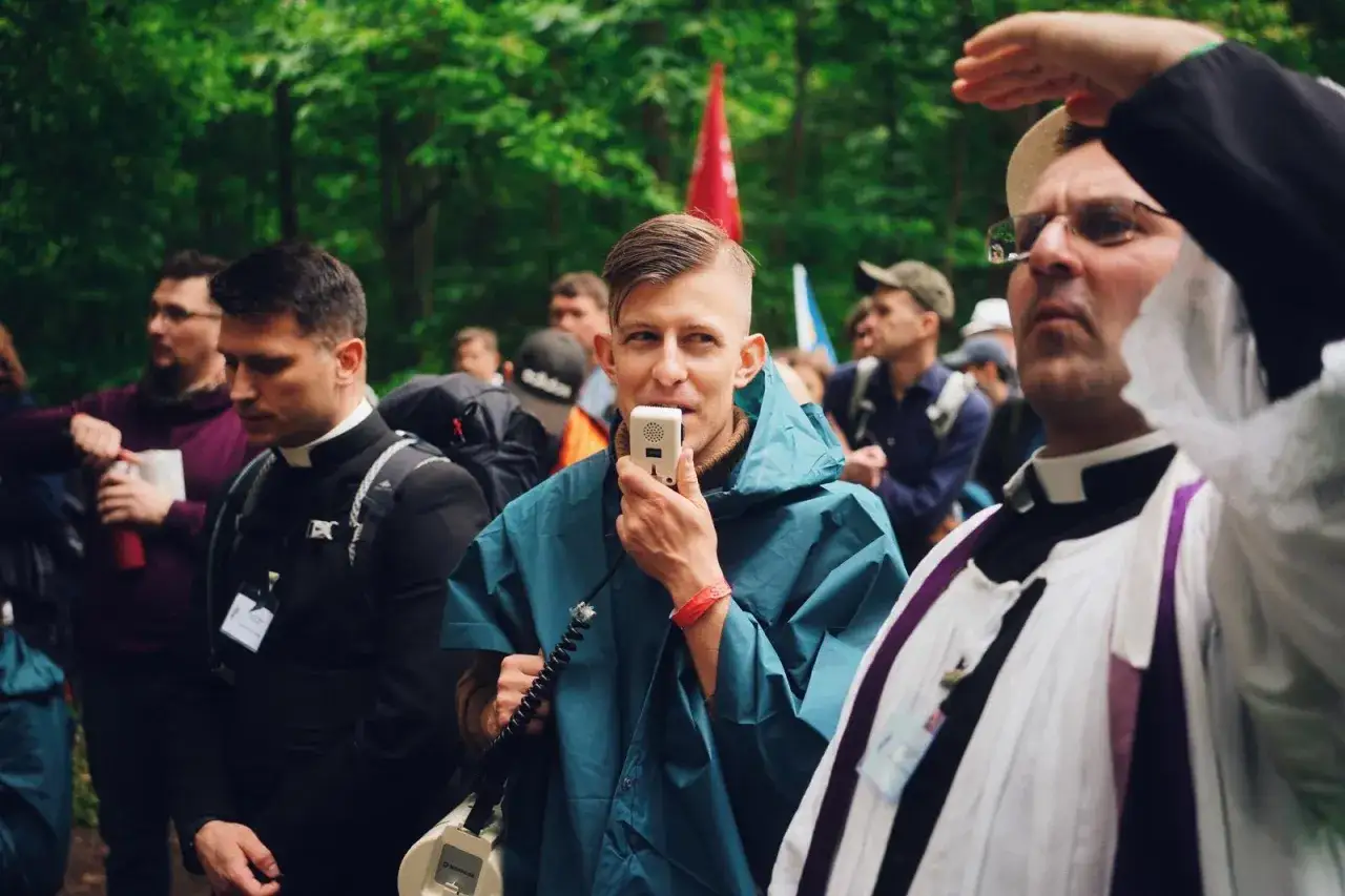 Mężczyzna w niebieskiej pelerynie mówi do mikrofonu, otoczony przez ludzi w strojach duchownych. Może to być fragment pielgrzymki, gdzie rozbrzmiewają pieśni pielgrzymkowe.