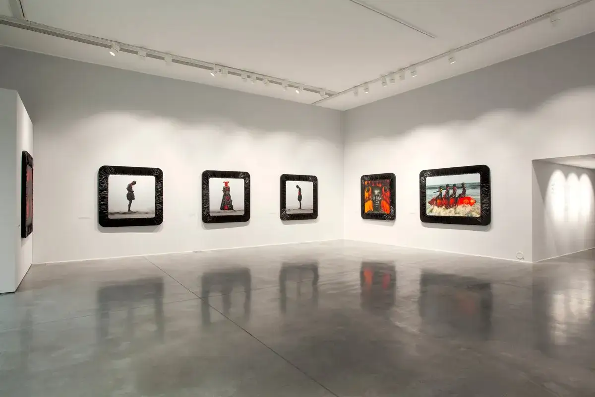 A modern gallery space showcasing striking fine art photography. The images, framed in black, depict diverse scenes, from solitary figures to groups on the water.