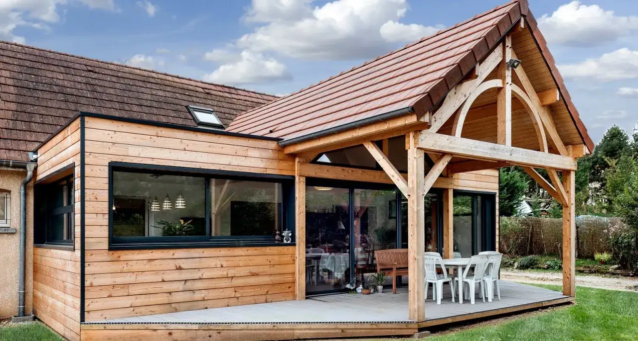 Belle extension de maison en bois avec une chambre lumineuse, une terrasse et une pergola.