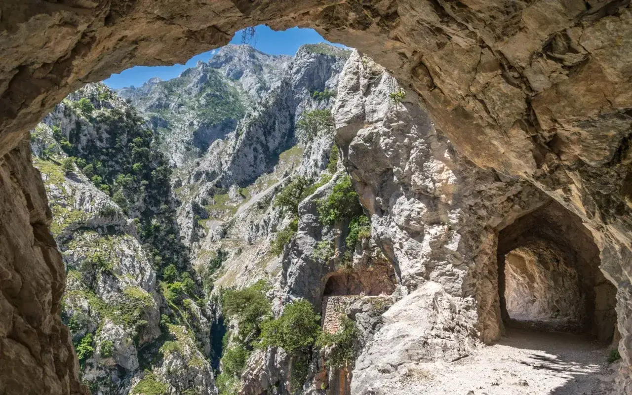 Blick durch einen Felsbogen auf die zerkl&uuml;fteten Berge der Picos de Europa. Ein schmaler Pfad f&uuml;hrt in einen dunklen Tunnel.