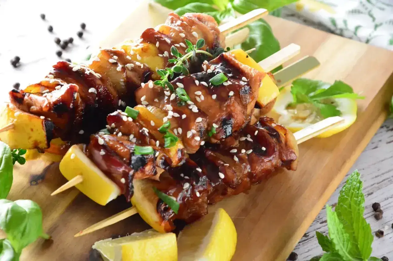 Aromatyczne yakitori na drewnianej desce, polane sosem, posypane sezamem i szczypiorkiem, z plasterkami cytryny i listkami mięty.