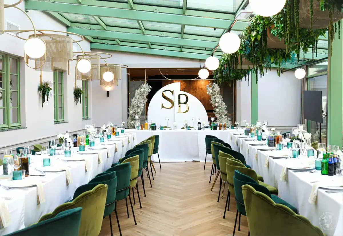 Elegancka sala weselna z długimi stołami, zielonymi krzesłami i dekoracjami z kwiatów. Monogram SB na tle podkreśla uroczysty charakter tego wyjątkowego slubu.