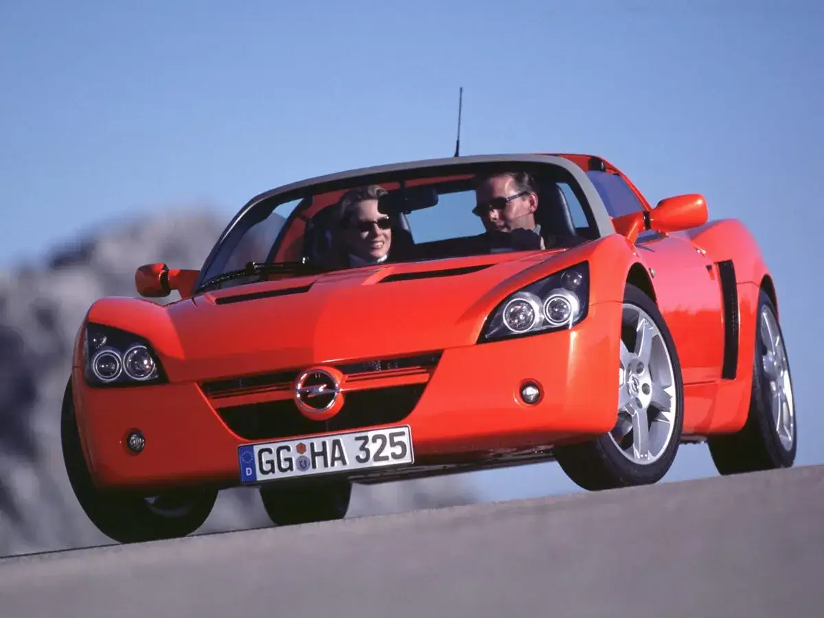 Pomarańczowy Opel Speedster z parą na pokładzie, cieszący się jazdą w słoneczny dzień.
