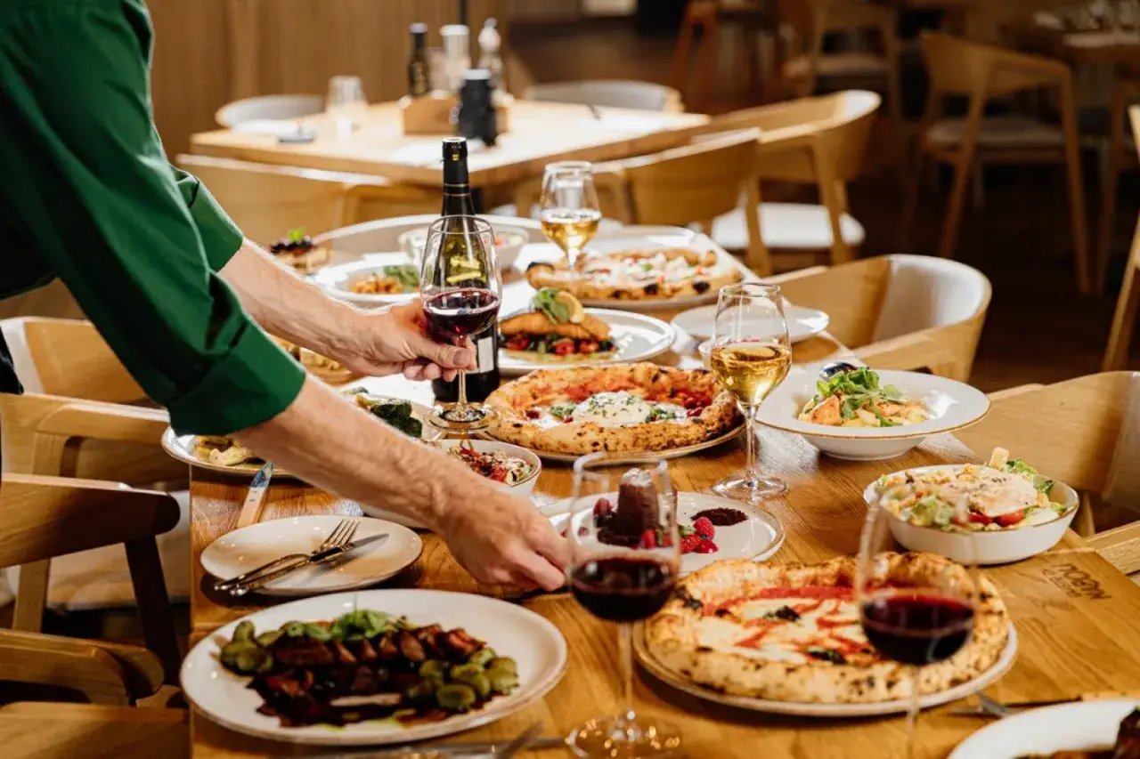 Włoska kolacja dla przyjaci&oacute;ł: stoły uginają się od pizzy, sałatek i wina. Kelner serwuje dania.