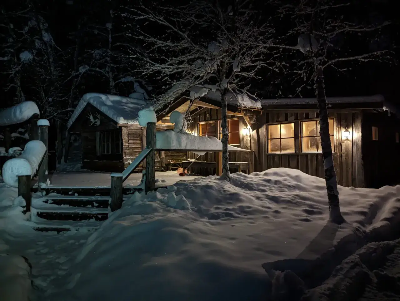 En snötäckt timmerstuga med upplysta fönster, en inbjudande svensk bastu i vinternatten.