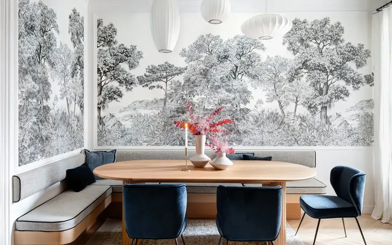 Un tableau panoramique noir et blanc orne le mur d'une salle à manger, représentant une forêt luxuriante. Une banquette grise et des chaises bleues complètent l'ensemble.