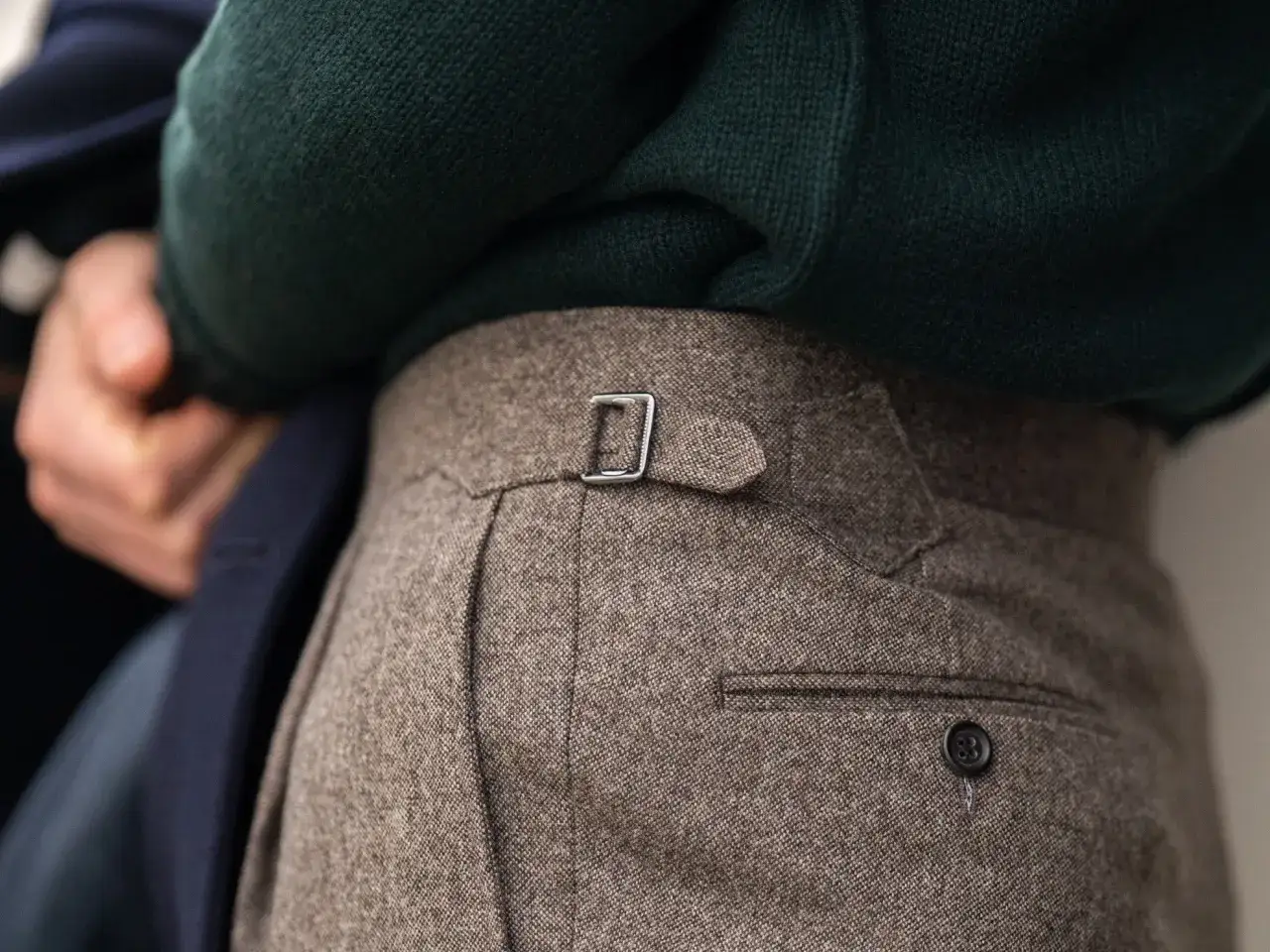 A man's tweed trousers with a silver buckle detail. He wears a dark green sweater and a navy jacket. Do you wear a belt with a suit?