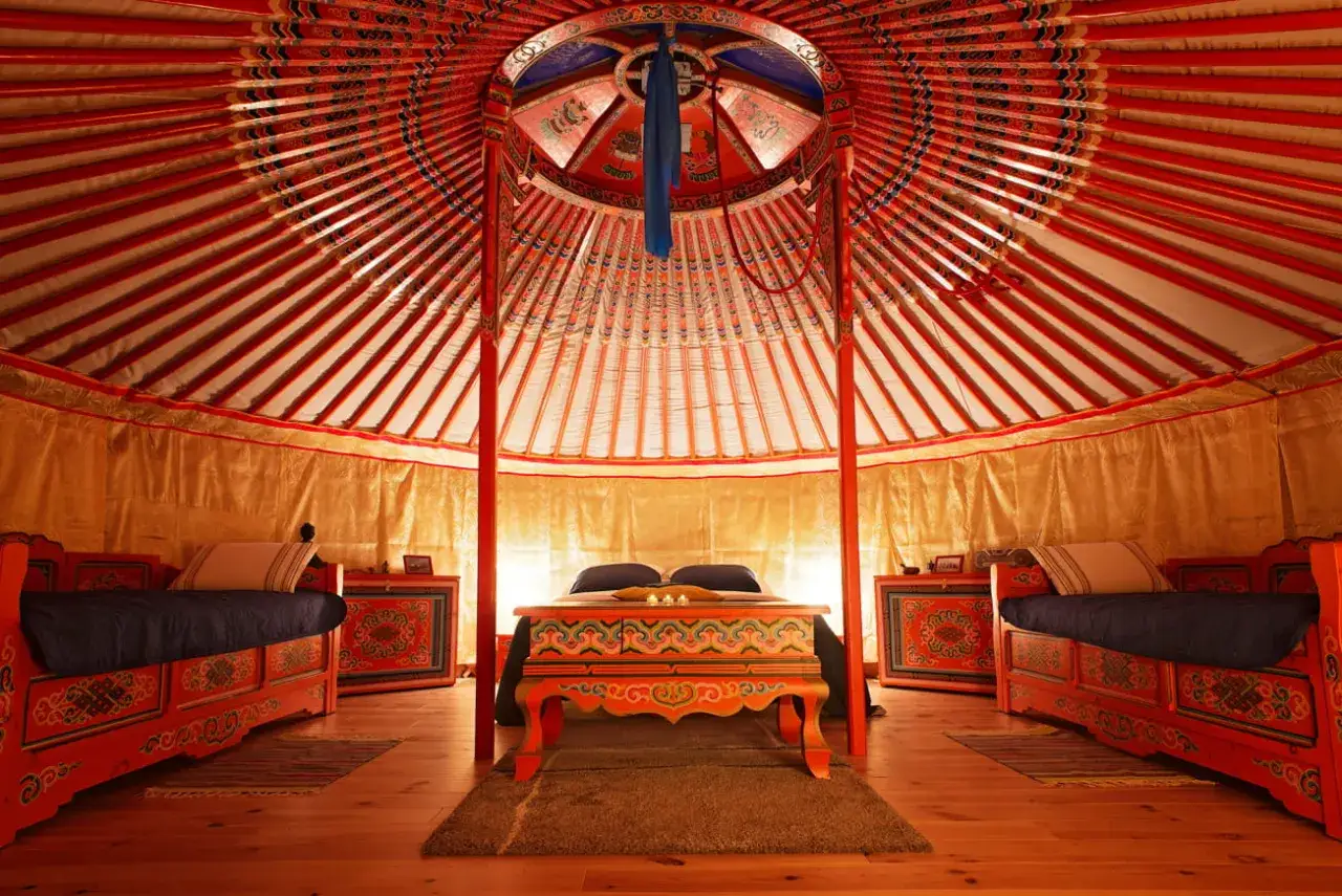 Intérieur d'une yourte alsacienne décorée de motifs colorés. Deux lits de chaque côté, un lit double au centre, et une table basse ornée.