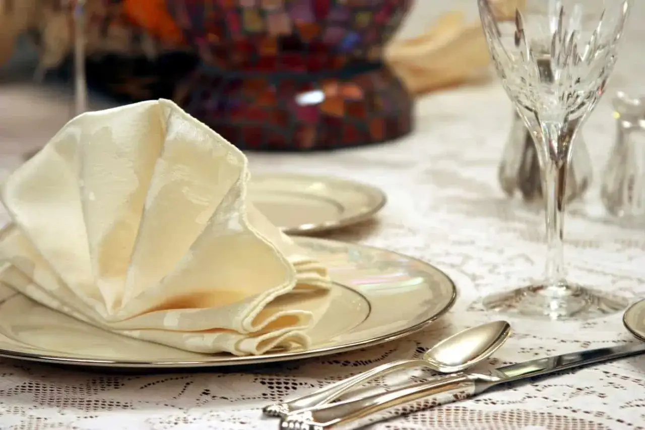 Un élégant **modèle serviette de table** en tissu crème, plié en éventail sur une assiette nacrée. La table est dressée avec des couverts en argent et un verre à pied.