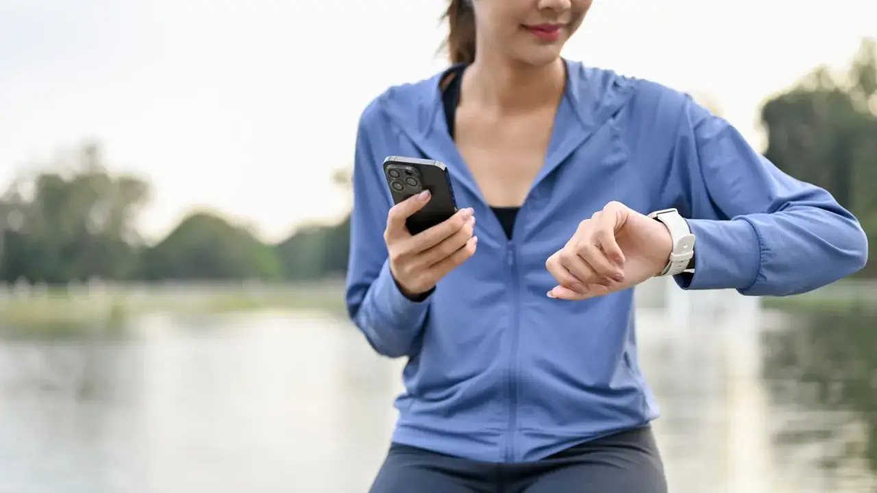 Kobieta w sportowym stroju sprawdza na zegarku aplikację do liczenia kroków, trzymając w drugiej ręce telefon.