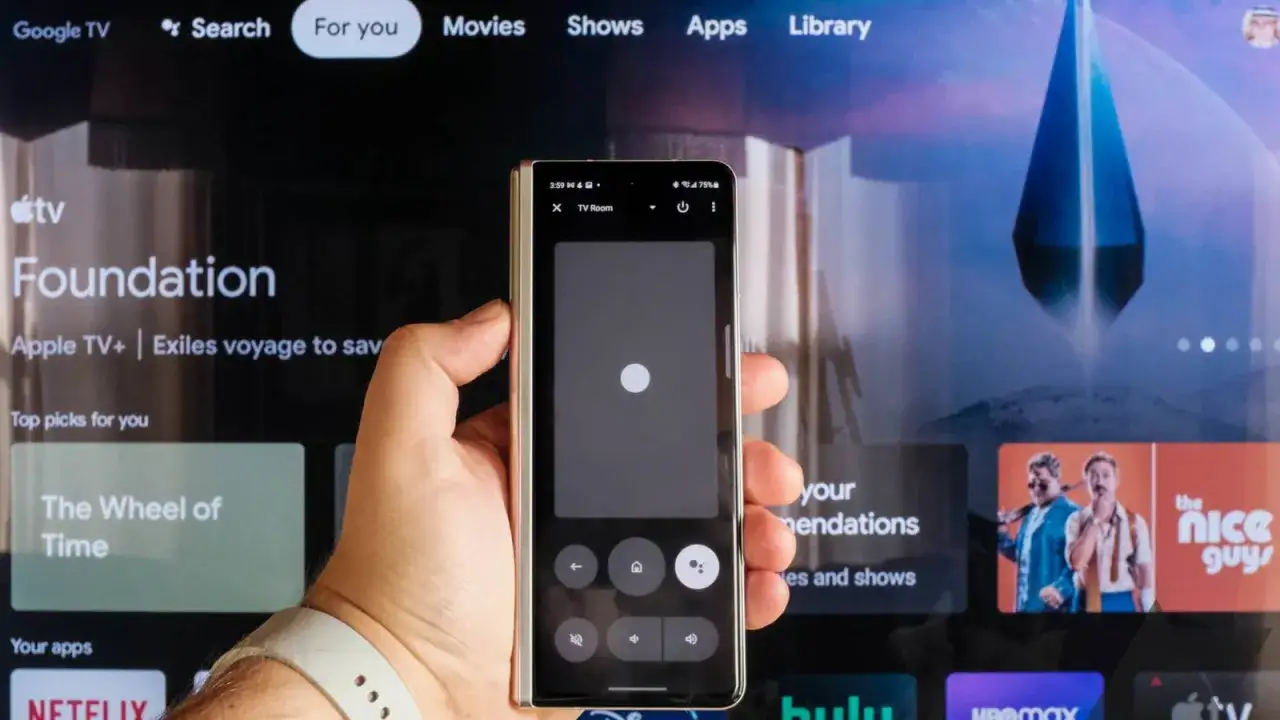 Ręka trzyma smartfon z aplikacją pilot do telewizora, sterując ekranem Google TV.