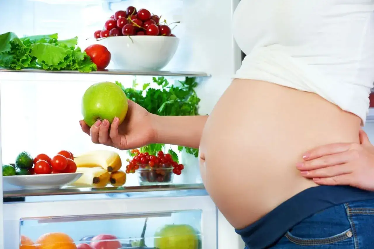 Gravid kvinna tar ett gr&ouml;nt &auml;pple ur kylen fylld med f&auml;rsk frukt och gr&ouml;nsaker.