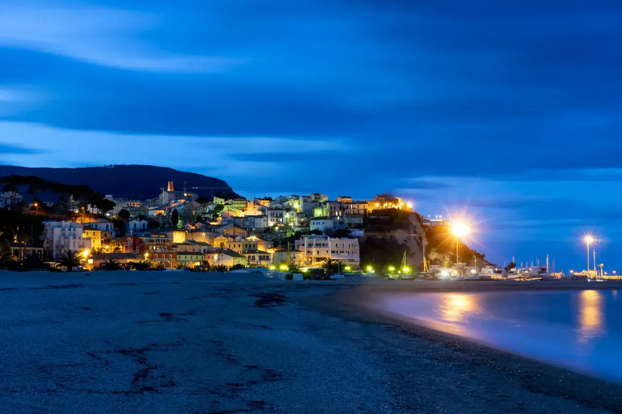 Il lungomare di Numana si illumina di notte, con le case colorate che si affacciano sul mare calmo e un porto pieno di barche.