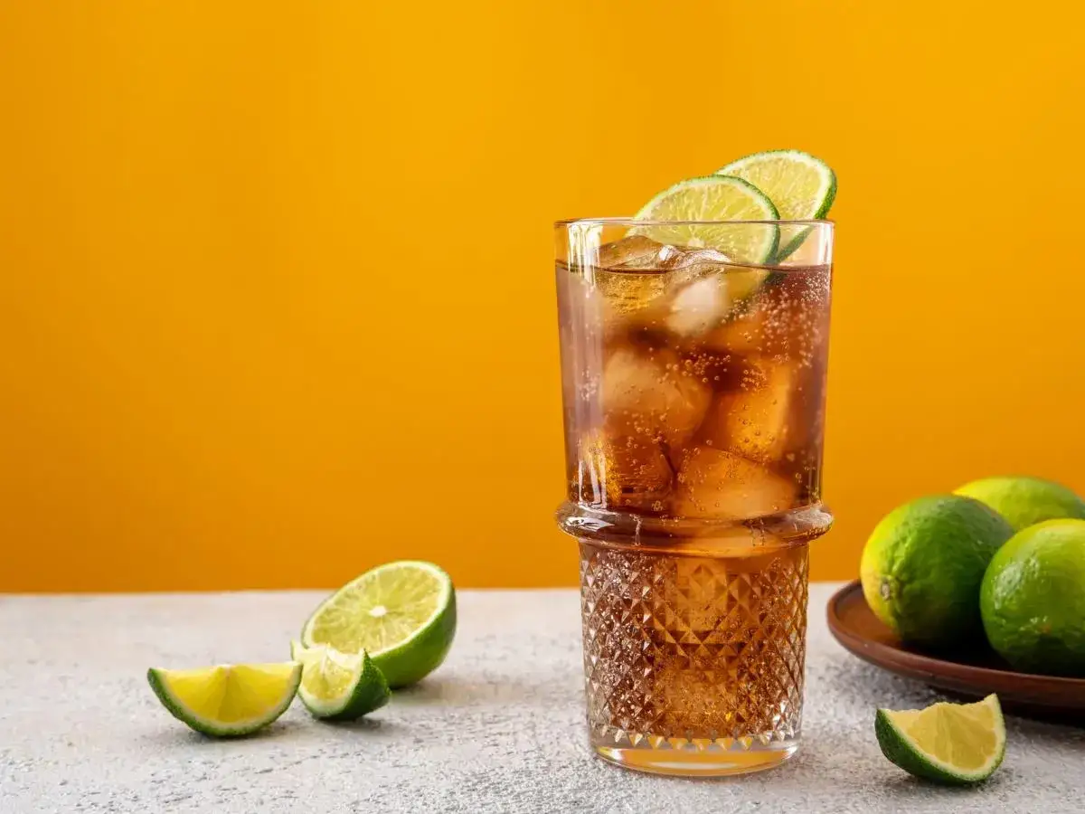 Idealne proporcje cuba libre: szklanka z lodem, rumem i colą, ozdobiona plasterkami limonki. Obok świeże limonki.