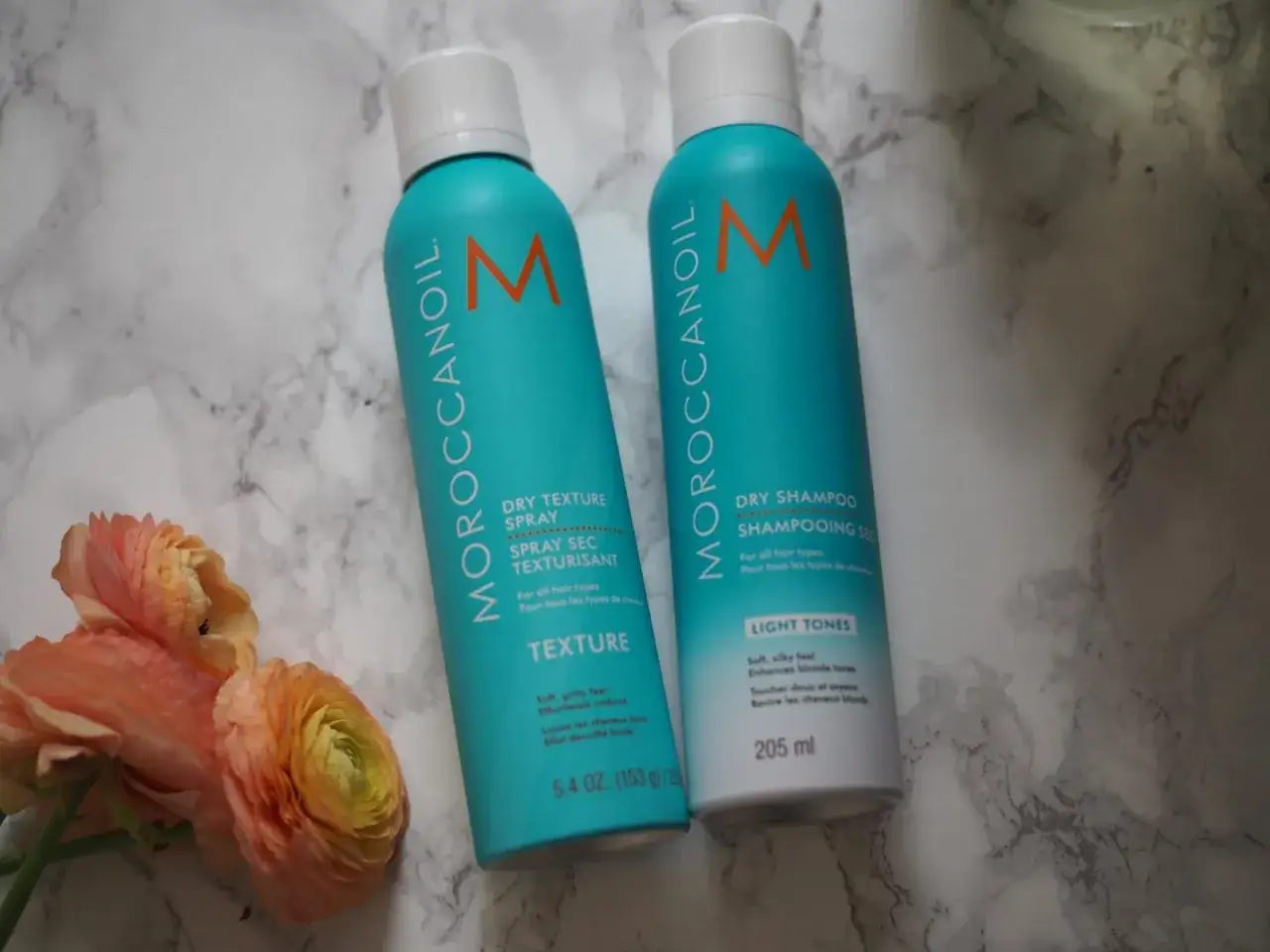 Dos aerosoles Moroccanoil, uno para texturizado de pelo y otro champú seco, junto a unas flores.