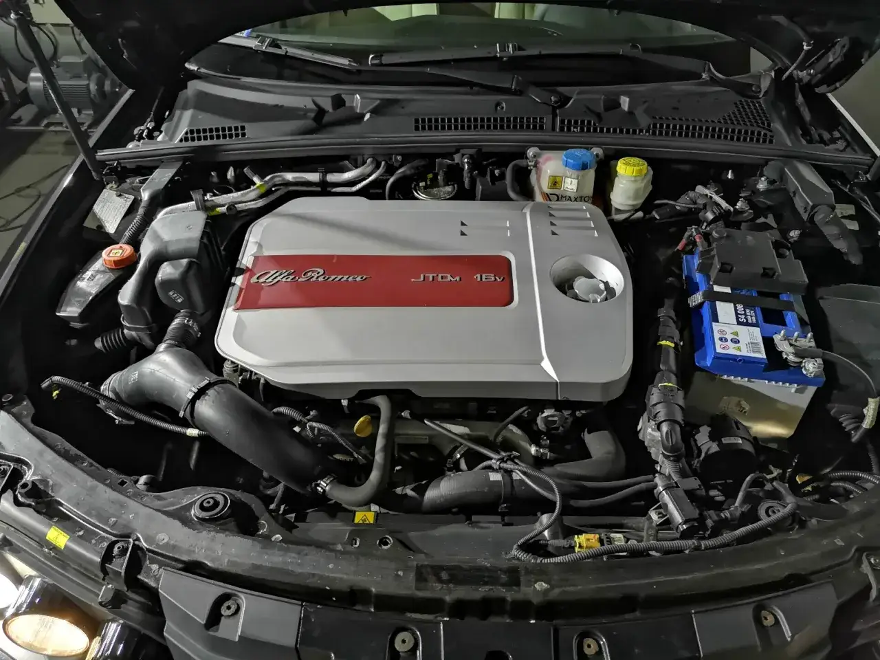 Silnik Alfa Romeo 159 1.9 JTD, z widocznym srebrno-czerwonym deklem i akumulatorem.