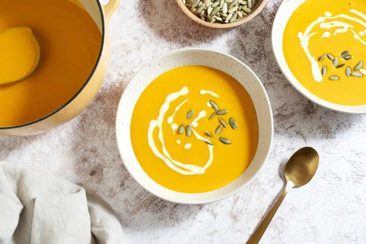 A creamy, vibrant kabocha squash soup is served in bowls, garnished with swirls of cream and pumpkin seeds. A pot of the soup and a spoon are also visible.