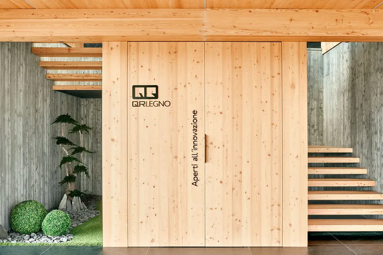 Porta scorrevole in **infissi in legno lamellare** con logo QR Legno. Scale in legno e cemento grezzo.