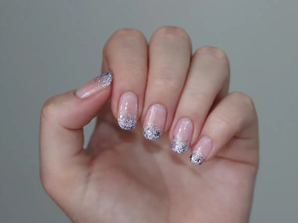 Dłoń z jasnymi paznokciami z brokatem, tworzącymi subtelny, błyszczący french manicure.