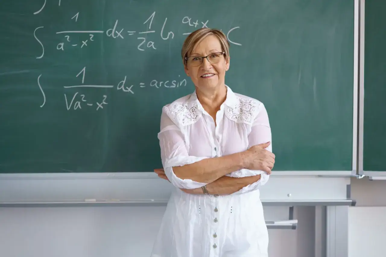 Eine Lehrerin vor einer Tafel mit Matheformeln. Sie l&auml;chelt und fragt sich: 