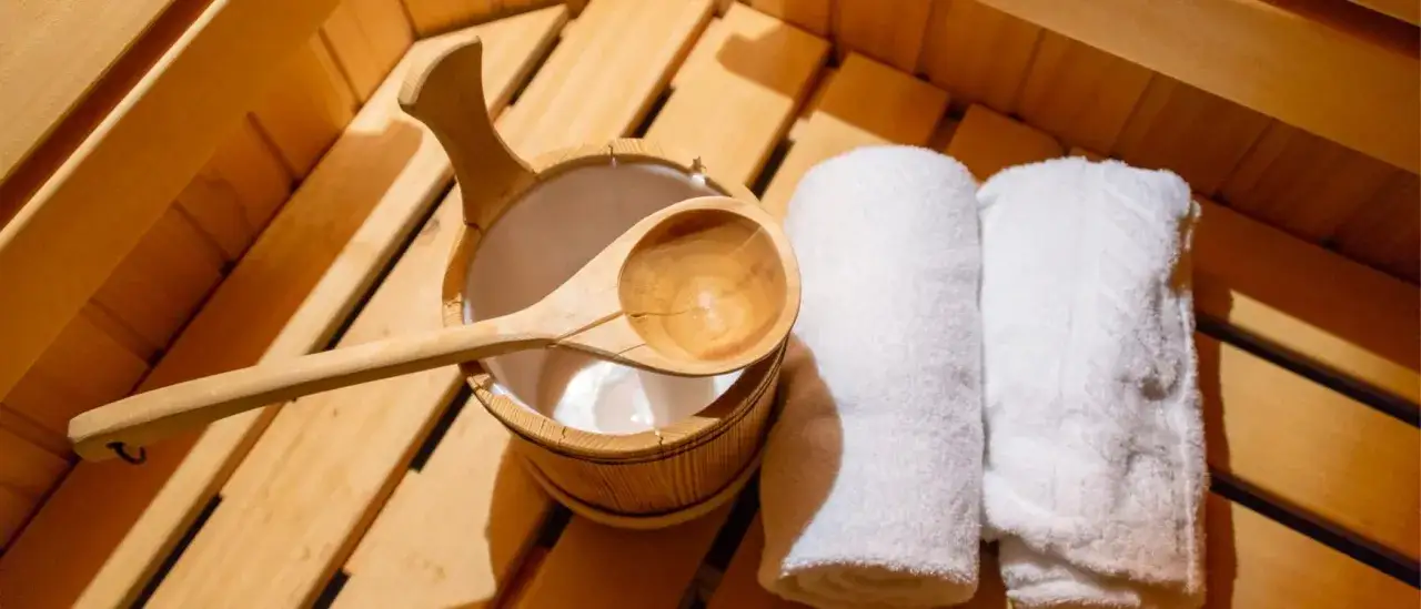 Secchiello e mestolo in legno, asciugamani bianchi arrotolati, pronti per un momento di relax. L'atmosfera &egrave; quella di una sauna, perfetta per chi cerca il benessere dei nudi in sauna.