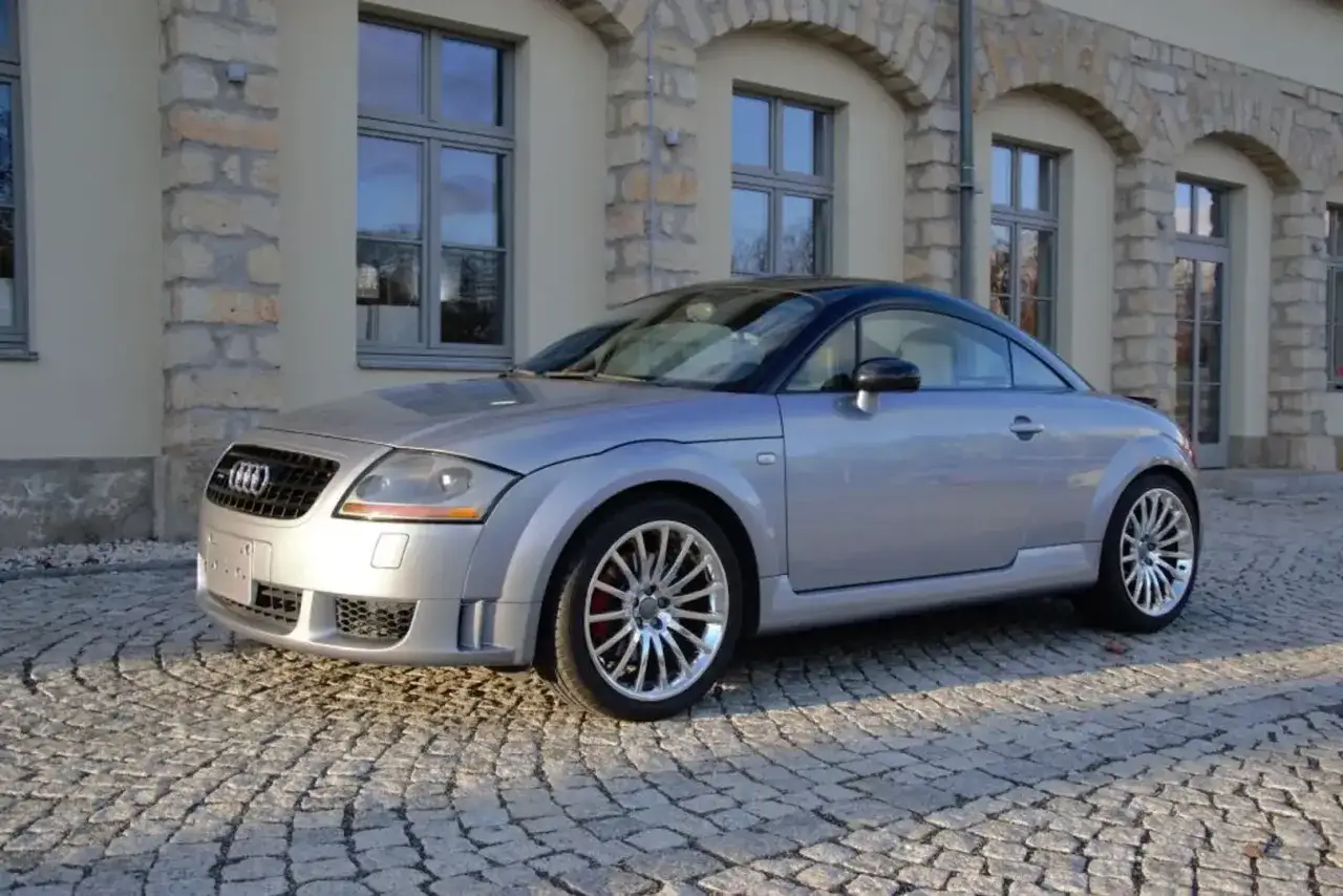 Silberner Audi TT 2005 mit schwarzem Dach parkt auf Kopfsteinpflaster vor einem Gebäude mit Steinbögen.