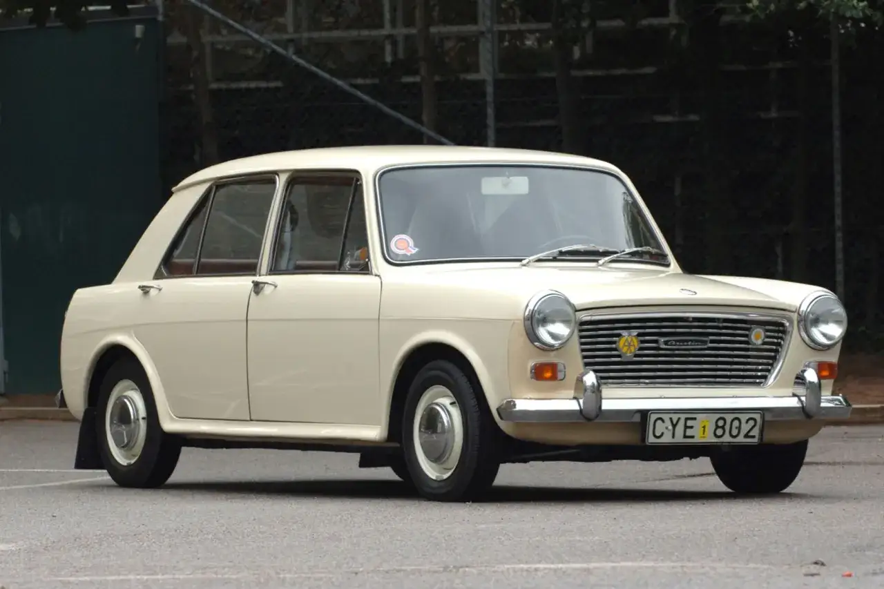 En krämvit Austin 1100 står parkerad på asfalt. Bilen har en klassisk design med kromade detaljer och en tydlig grill.