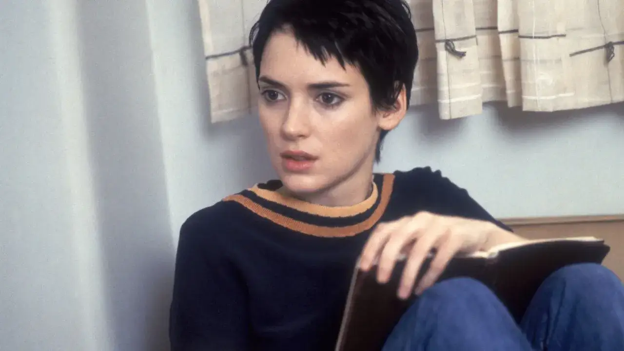Winona Ryder joven, con pelo corto y suéter azul con rayas, lee un libro.